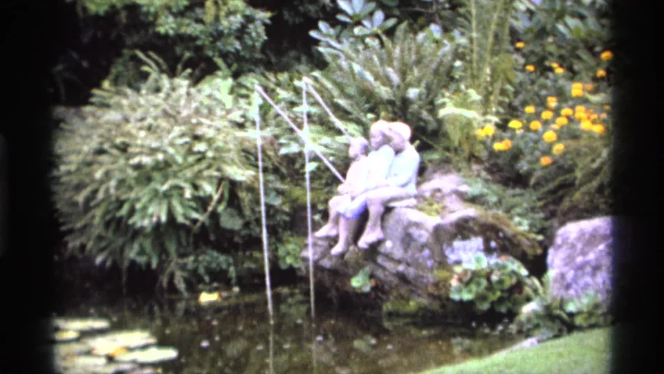 1974 Vancouver garden view showing a fishing-figure statue on rocks beside a pond, with water streams, flowers, and surrounding greenery.