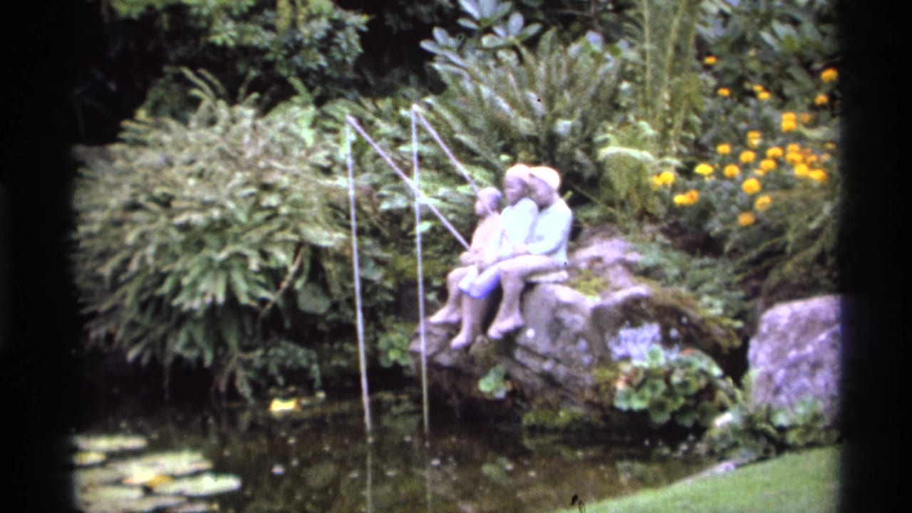 1974 Vancouver 8mm frame showing a two-child fishing statue on mossy rocks by a garden pond with ferns, lily pads, and yellow flowers.