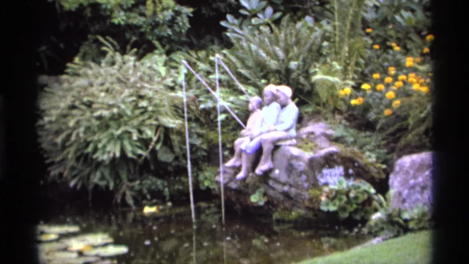 1974 Vancouver garden view with a decorative child fishing statue on rocks above pond water, surrounded by dense planting and yellow flowers.