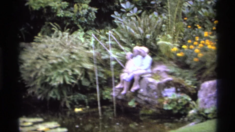 Soft 1974 Vancouver 8mm frame of the fishing statue on rocks beside a pond, with lily pads, ferns, and yellow flowers in view.