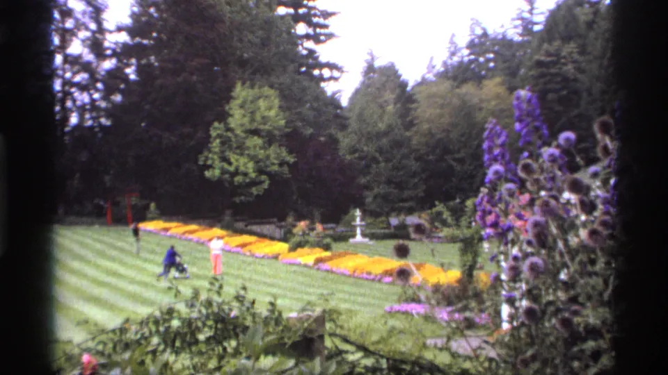 1974 Vancouver garden frame showing visitors on a striped lawn, colorful flowerbeds, a white fountain, tall trees, and purple flowers in the foreground.