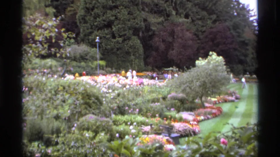1974 Vancouver 8mm frame of planted garden beds, clipped shrubs, a lawn edge, mature trees, and distant visitors in a public park.