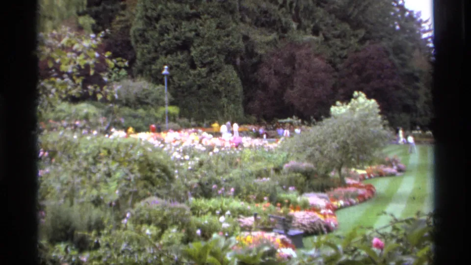 1974 Vancouver garden frame with bright flower beds, shrubs, lawn, trees, and distant visitors rendered in soft 8mm color.