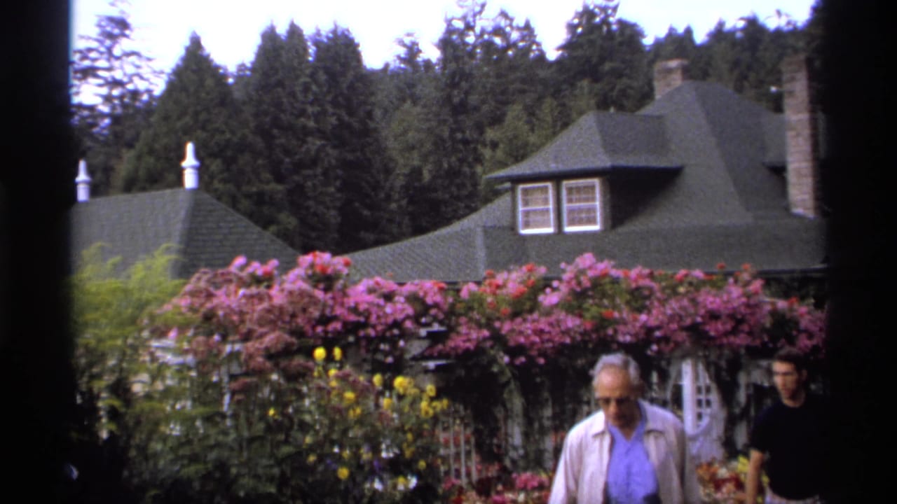 Garden building with green roof behind pink climbing roses as adults pass nearby.