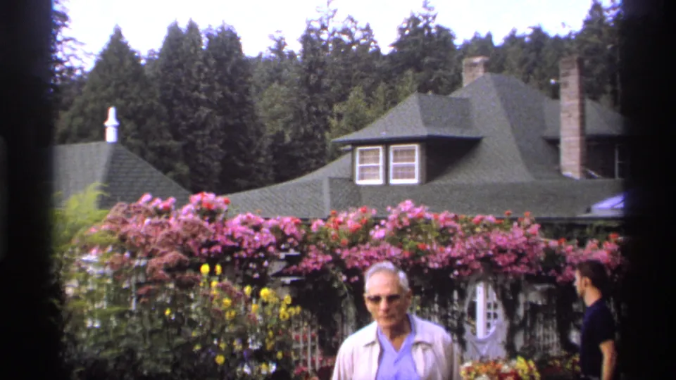 Adult man in sunglasses walks through a 1974 Vancouver garden, with pink climbing roses, flowers, trees, and a green-roofed building behind him.