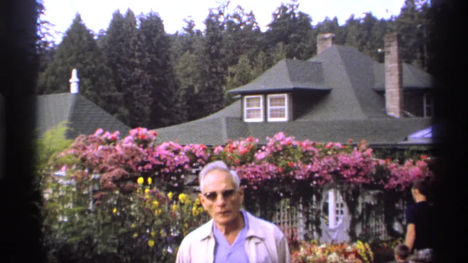 In the 1974 Vancouver garden footage, an adult man stands near the camera before pink rose trellises, yellow flowers, trees, and a green-roofed building.