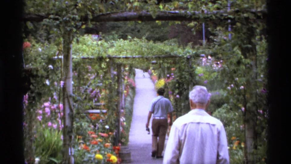1974 Vancouver home-movie frame of two adult visitors walking through a leafy arbor-lined garden path.