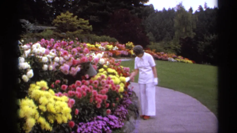 Adult woman in white clothing walks along a curved garden path beside dense pink, yellow, purple, and white flowers in Vancouver.