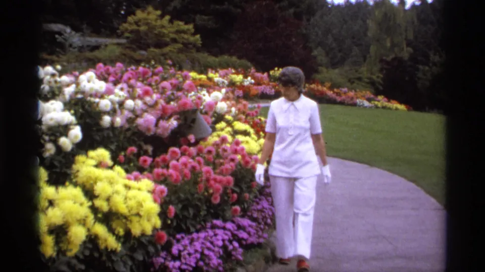 A woman in white appears on a Vancouver garden path in 1974, looking toward rows of bright flowers and lawn.