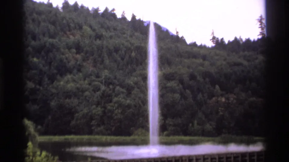 A high water fountain rises above a pond before a dense forested hillside in Vancouver archival footage.