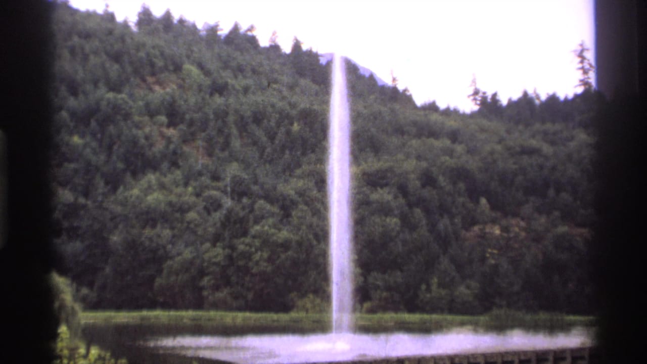 Tall fountain spraying from a lake with forested hillside behind it and dark window framing in front.
