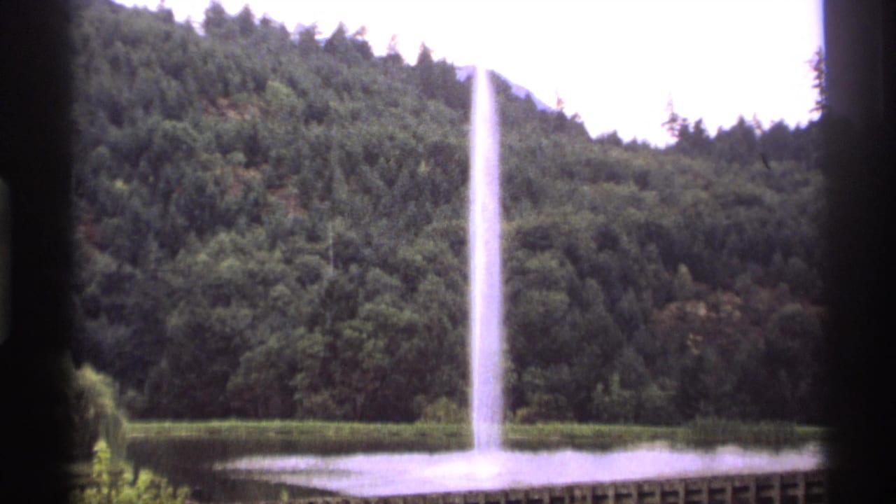 Lake fountain and forested hillside seen through dark vertical and side window framing.