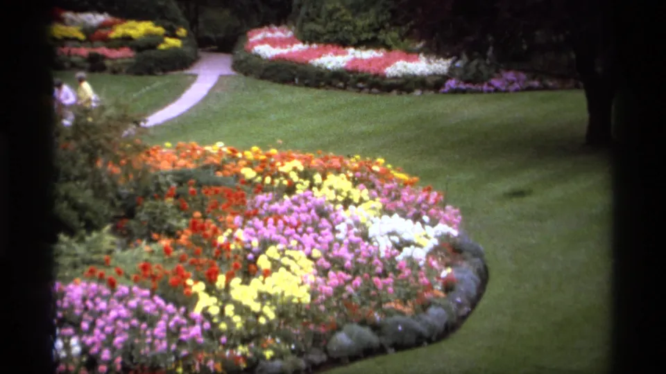 1974 Vancouver home-movie frame with bright flower beds, manicured grass, winding paths, shrubs, and distant garden visitors.