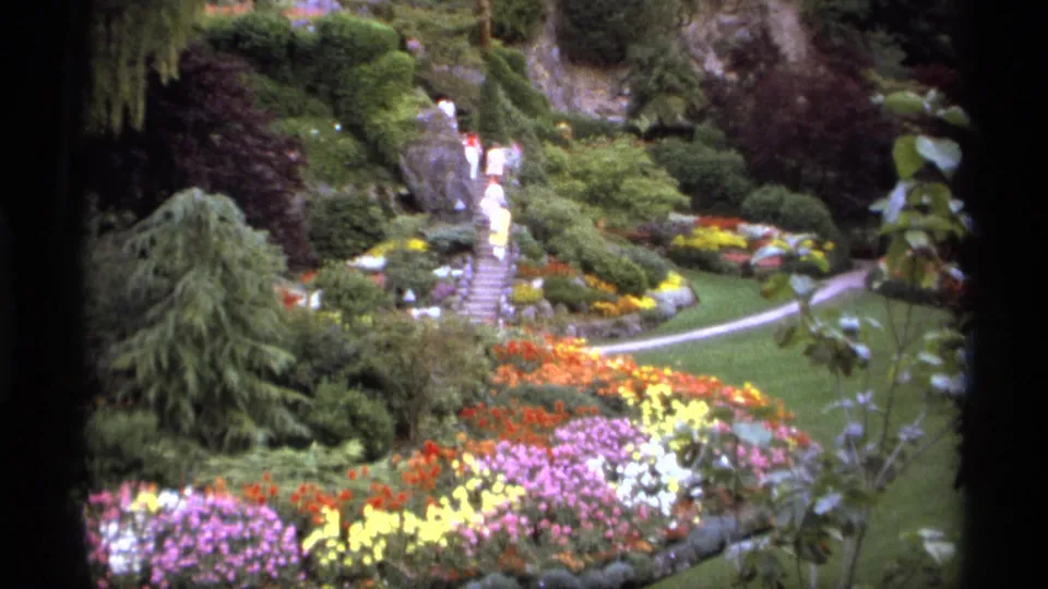 In a Vancouver garden, visitors in light clothing move near central stone steps above colorful beds, shrubs, lawn, and a curved path.