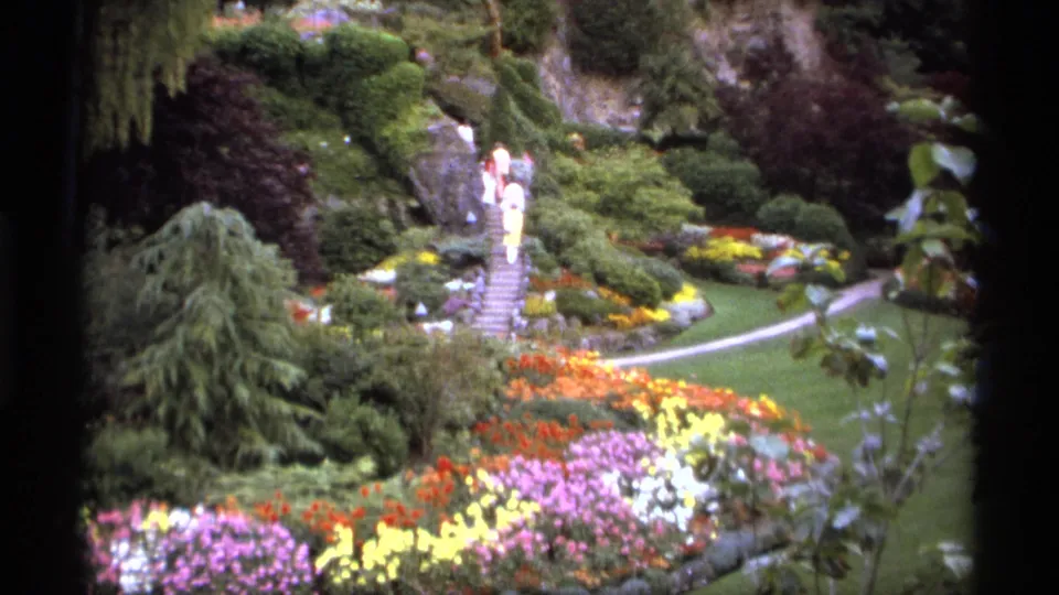 1974 Vancouver home-movie frame showing visitors on garden steps, dense shrubs, bright flower beds, a lawn, and a curved walkway.