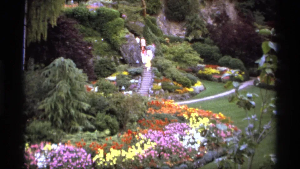 Vancouver garden footage from 1974 with visitors on central steps, colorful flower beds, green lawn, shrubs, and a winding pale path.
