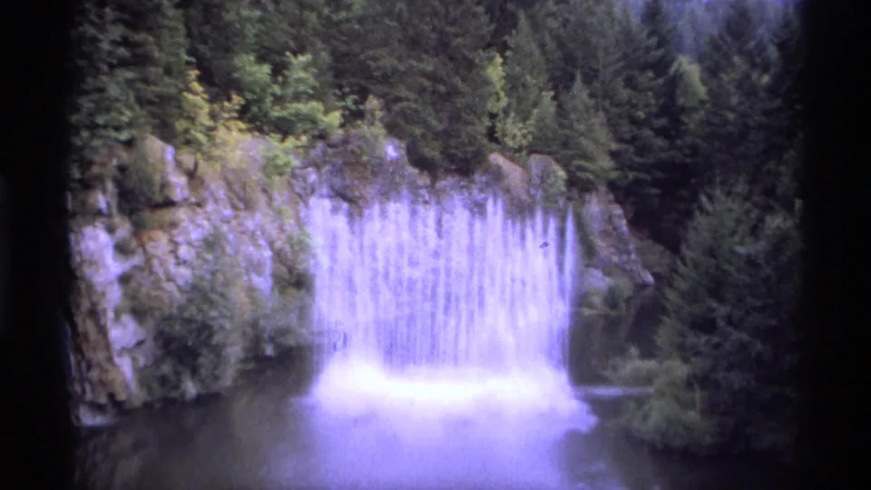 1974 Vancouver home-movie frame showing a wide waterfall, rocky banks, dense evergreens, and a still pool below.