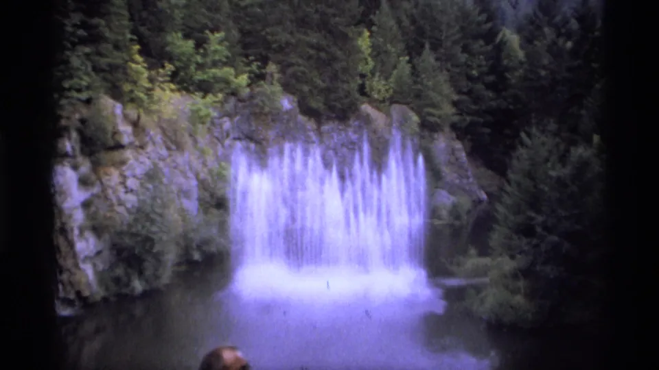 1974 Vancouver home-movie frame of a waterfall and surrounding trees, with a small visitor partly visible near the water.