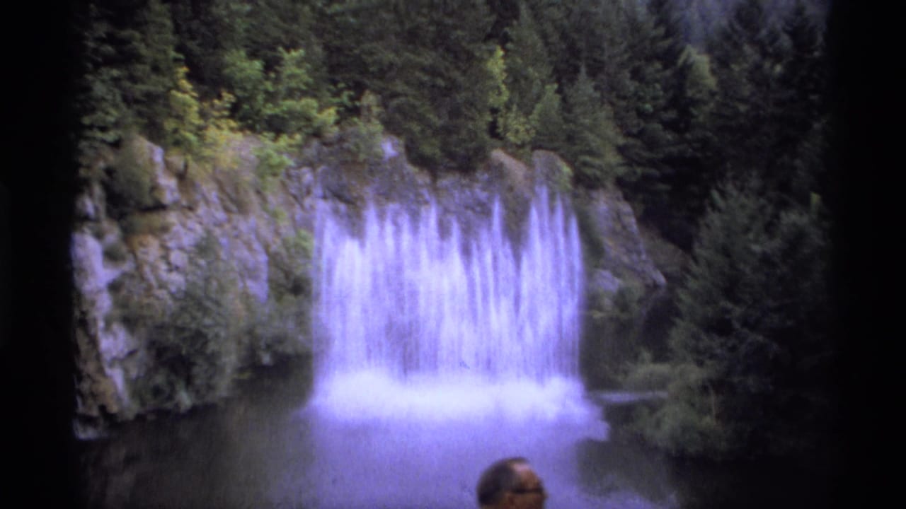 1974 Vancouver-area 8mm frame showing broad water jets in a pool beneath rock and evergreen trees, with a partial head at the lower edge.