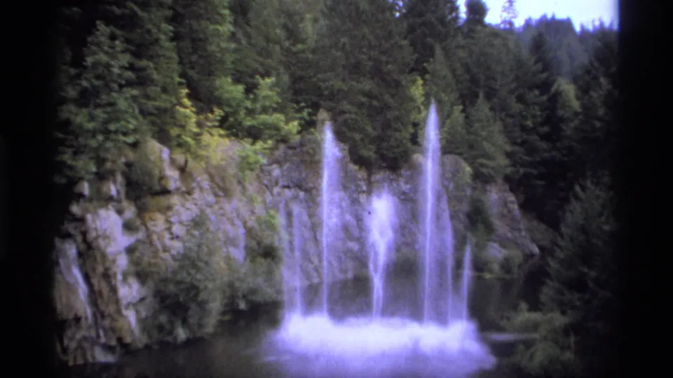 1974 8mm travel frame near Vancouver showing tall water plumes, mist, rock faces, and dense evergreen forest in daylight.