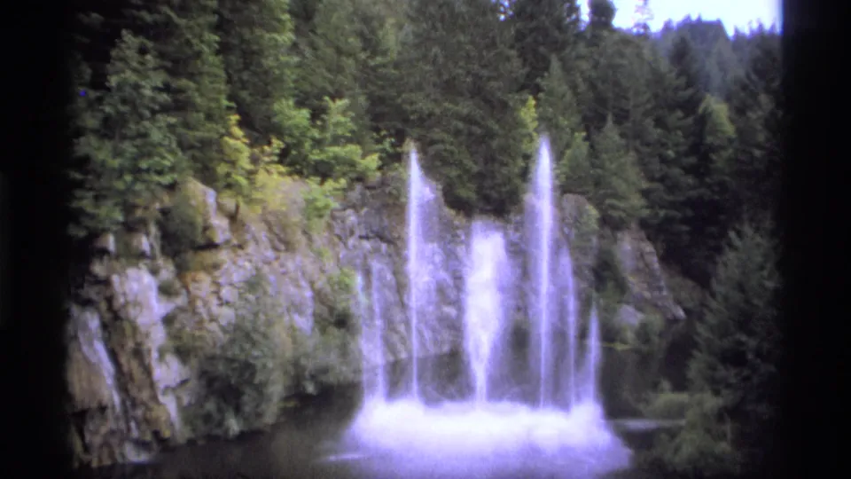 1974 Vancouver-area home-movie frame with multiple bright water jets, a rocky bank, dark pool water, and thick conifer trees.