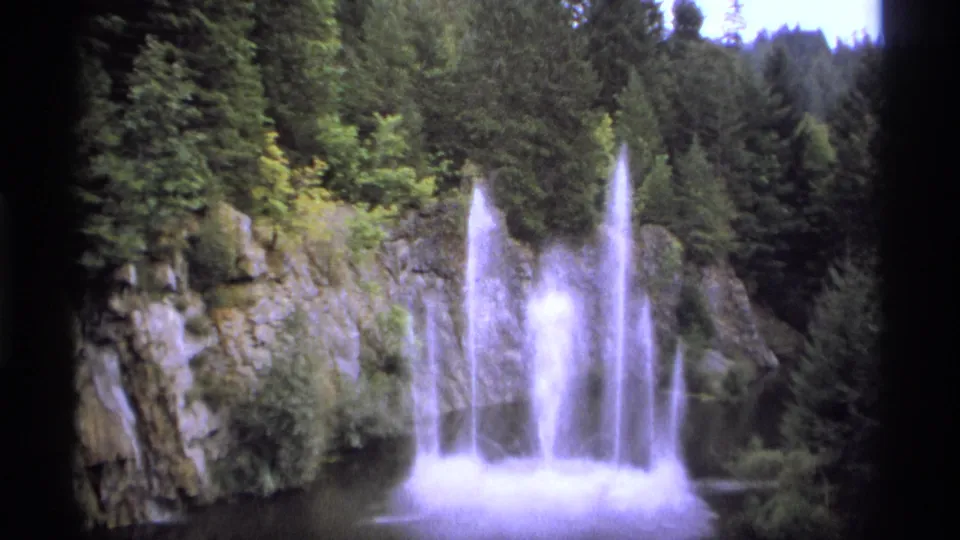 1974 8mm frame near Vancouver showing several water columns erupting from a pool with rock, shrubs, and evergreen trees behind them.
