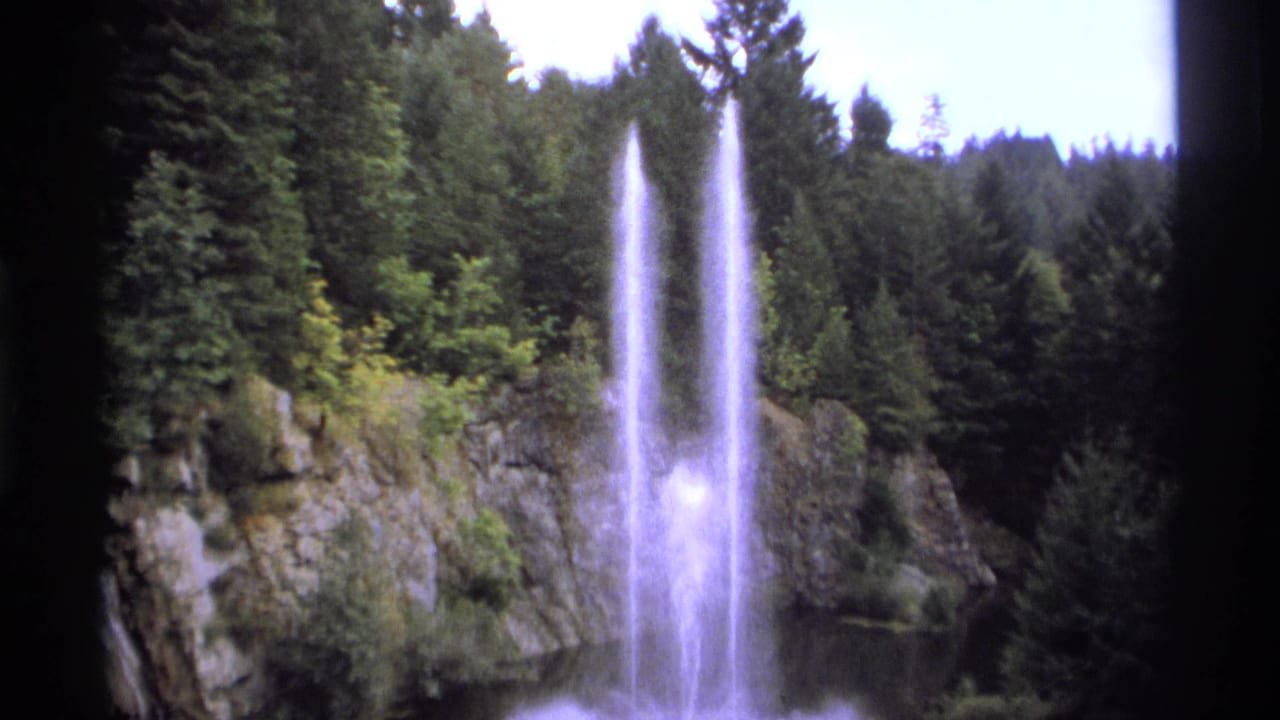 1974 Vancouver-area 8mm frame showing two tall water plumes against rocky cliffs and dense conifer forest.