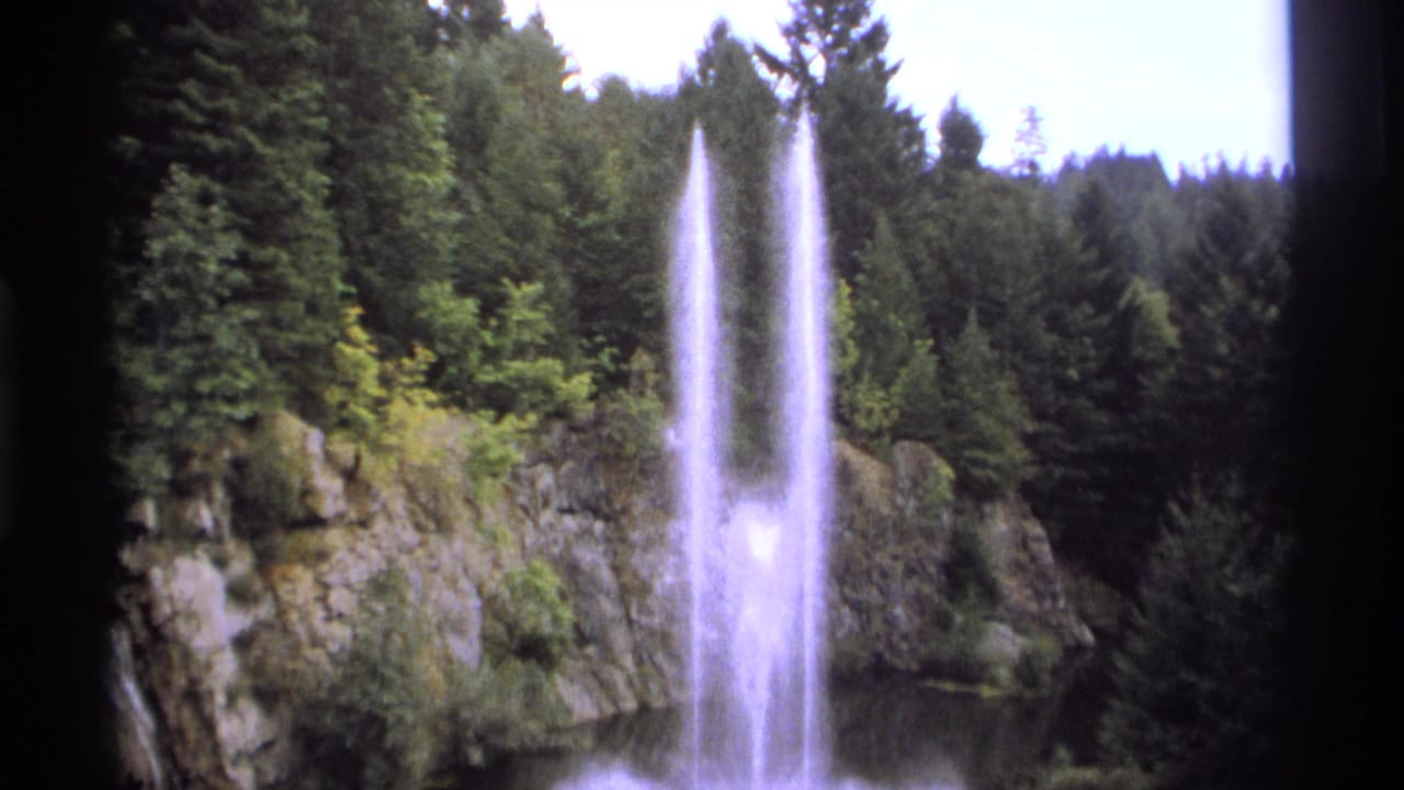 Tall fountain spray stands before a rocky cliff and dark evergreen forest.