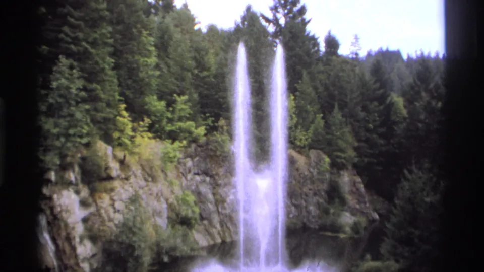 1974 Vancouver-area 8mm frame showing twin fountain plumes, mist, cliff rock, and conifer trees from a visitor viewpoint.