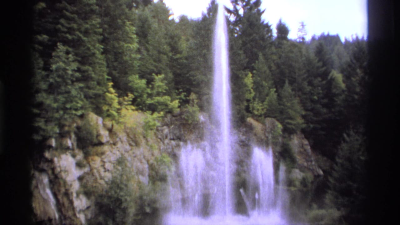 Tall fountain jet rising beside rocky cliffs and evergreen trees.