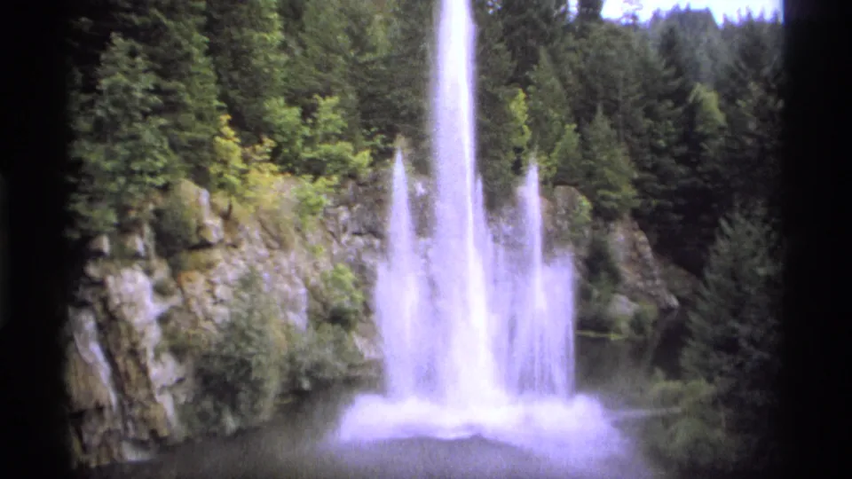 The 1974 8mm frame shows the fountain at full spray, with white water blooming above a pond in a rocky evergreen setting.