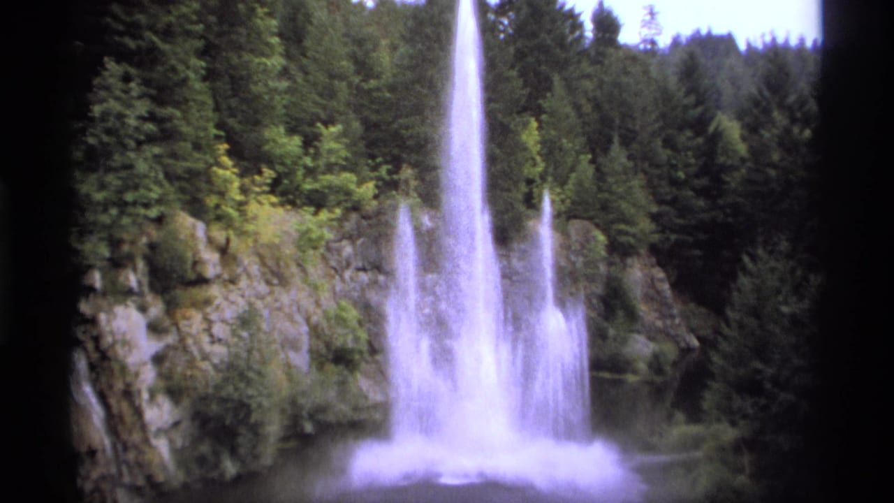 1974 Vancouver home-movie frame showing a tall water plume or waterfall spray against rocks and dense evergreens.