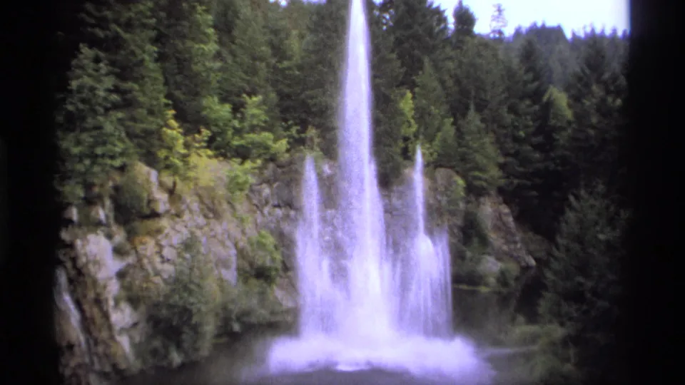 1974 Vancouver home-movie frame showing a tall water plume or waterfall spray against rocks and dense evergreens.