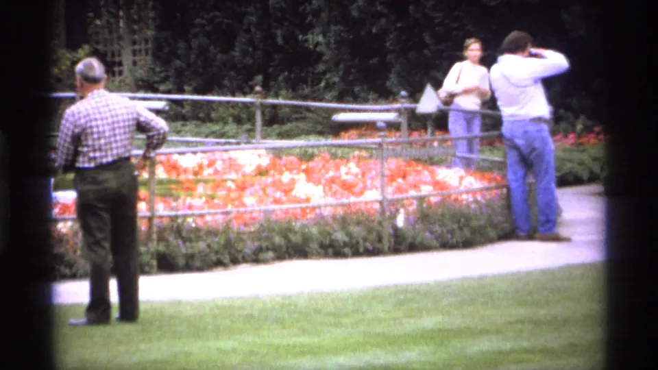Adults in a Vancouver park garden stand beside a fenced flower bed, with one visitor appearing to take a photograph.