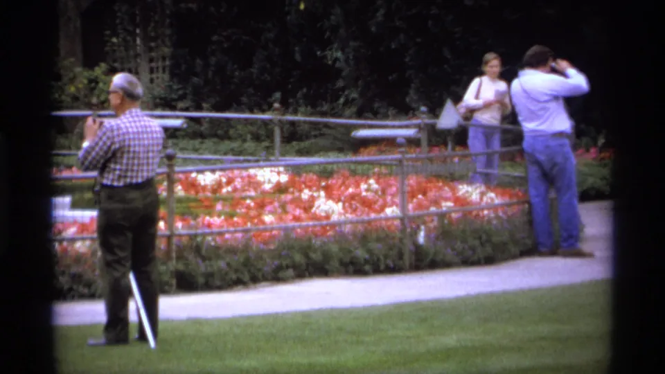 Three adults remain beside a fenced flower bed in 1974 Vancouver, including a man with a white cane and another with a camera.
