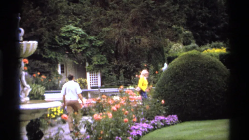 1974 Vancouver garden frame with adults walking near blooming beds, manicured shrubs, a lawn edge, and a tiered fountain partly in view.