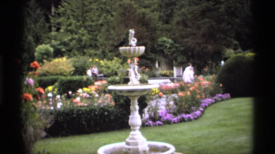 1974 Vancouver garden frame centered on a tiered fountain with surrounding flowerbeds, trimmed hedges, lawn, and adult visitors in the background.