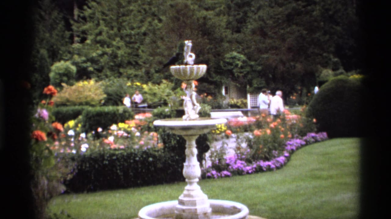 1974 Vancouver home-movie frame showing a white tiered fountain, colorful flower beds, trimmed hedges, lawn, trees, and distant visitors.