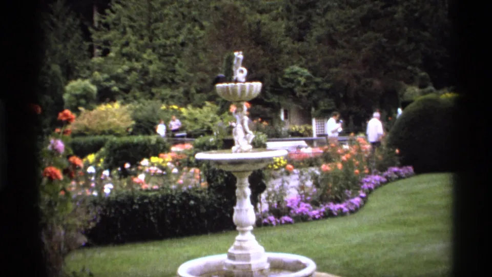 1974 Vancouver garden view with a white fountain centered among colorful beds, clipped hedges, lawn, benches, trees, and visitors in the distance.