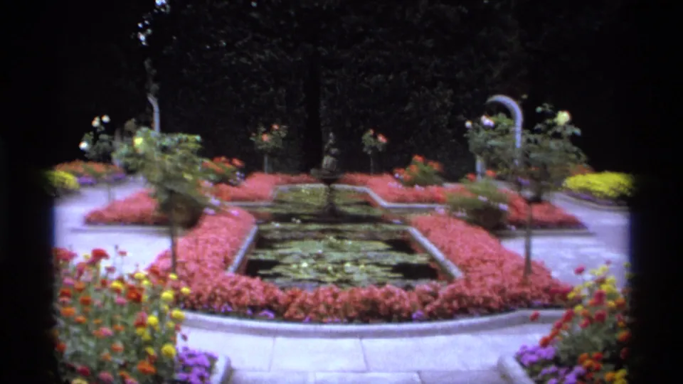 1974 Vancouver 8mm frame of a landscaped garden, with a rectangular pond, pink flower beds, small trees, paths, and vivid foreground blooms.