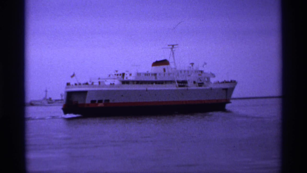 1974 8mm travel footage shows a large white ferry with a red hull moving across calm harbor water, with small distant figures likely on deck.
