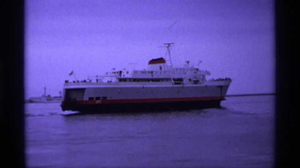 1974 8mm travel footage shows a large white ferry with a red hull moving across calm harbor water, with small distant figures likely on deck.