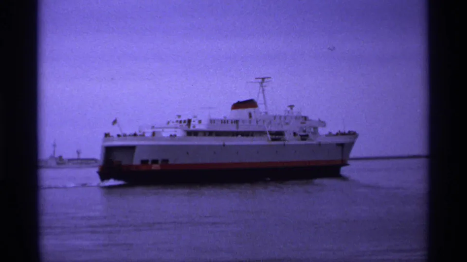 A large ferry continues across calm water in the 1974 home-movie footage, framed by dark film edges and a hazy shoreline.