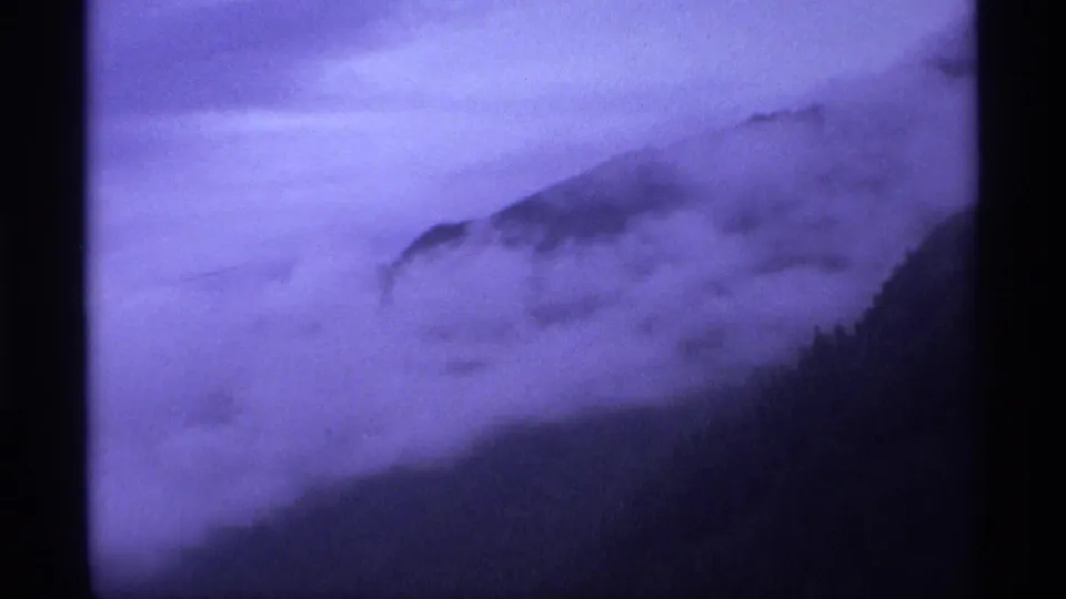 1974 home-movie frame cutting from city signage to a misty mountainside, with low cloud or fog obscuring the slope.