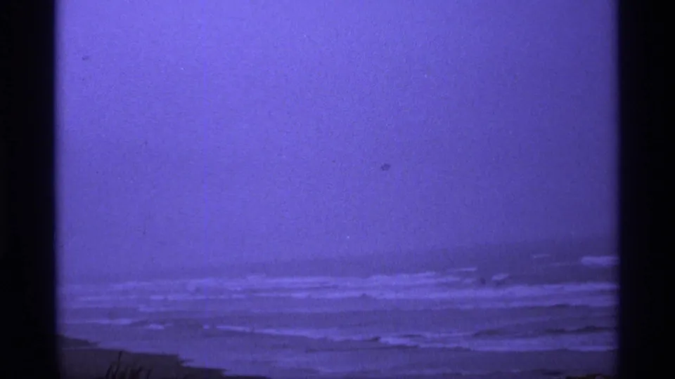 Purple-toned 8mm coastal view with low surf lines, hazy horizon, and a dark shoreline edge, supported by Vancouver clip metadata.