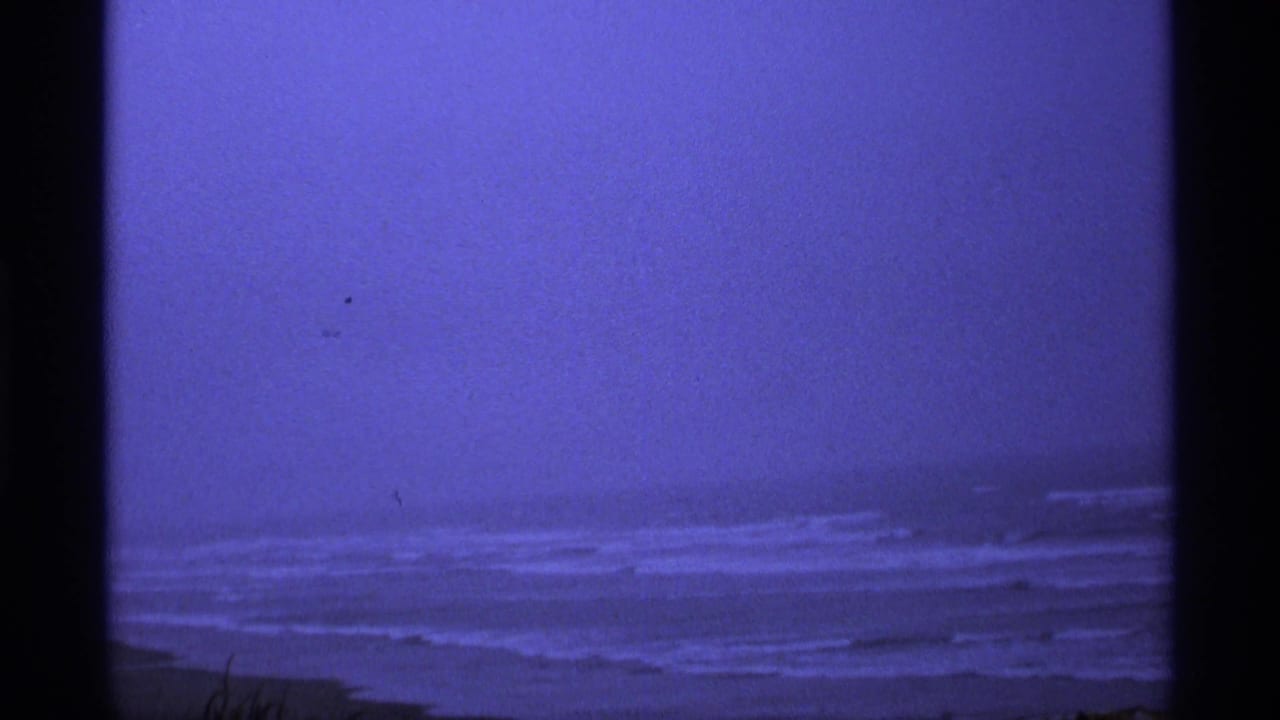 1974 Vancouver-area 8mm frame with blue-tinted surf rolling toward a dark beach under a hazy sky.