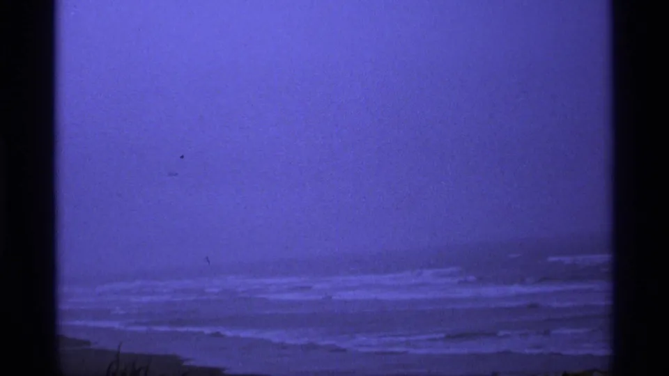 1974 Vancouver-area 8mm frame with blue-tinted surf rolling toward a dark beach under a hazy sky.