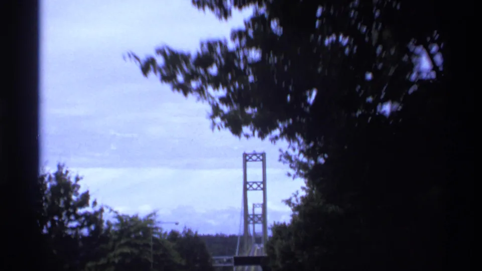 1974 Vancouver-area car-window view of a suspension bridge tower framed by dark trees and pale cloudy sky.