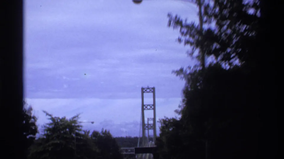 1974 Vancouver home-movie frame showing a suspension bridge tower partly framed by dark trees and roadside shadows.
