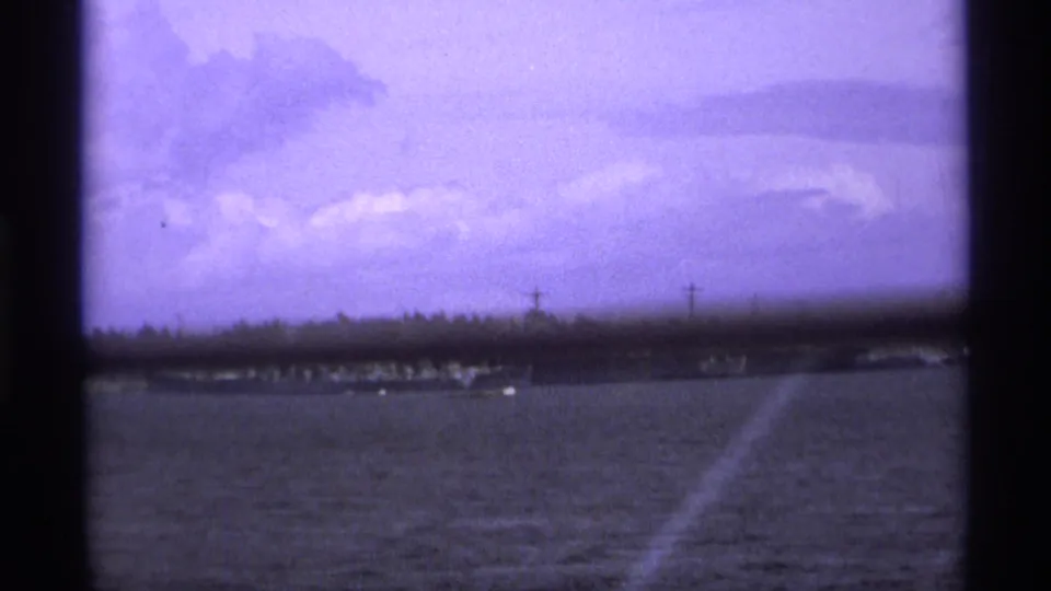 1974 Vancouver home-movie frame showing a distant military ship near a low shoreline under a purple-tinted cloudy sky.